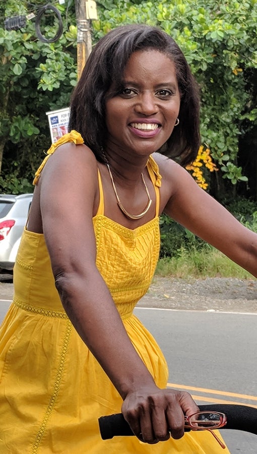 woman wearing yellow spaghetti-strap dress on bike on road, trees and car in background