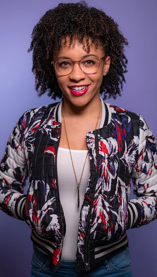 Leah wears her hair naturally curly, a multi-colored jacket, long gold necklace, white t-shirt and jeans, and stands against a purple backdrop.