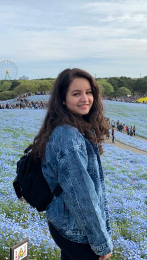 Young woman in denim jacket stands in field of small flowering blue flowers; lines of people, trees and a ferris wheel are visible in the background