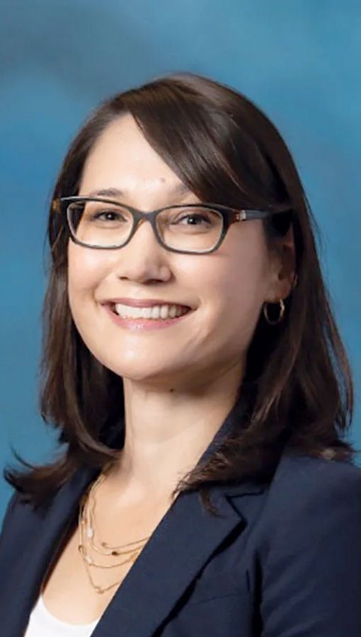 Woman with dark straight hair, dark-rimmed glasses, navy blue blazer, white shirt and layered necklaces smiles at camera in front of teal background