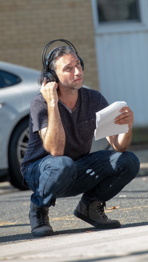 Man with shoulder-length silver hair, wearing jeans and dark t-shirt, crouches on pavement and holds earphones on head while looking in front of/above him