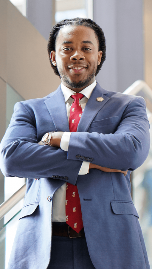 professional portrait of young Black man in blue-grey suit, white button-down shirt, red tie, standing in sunny stairwell, with arms crossed