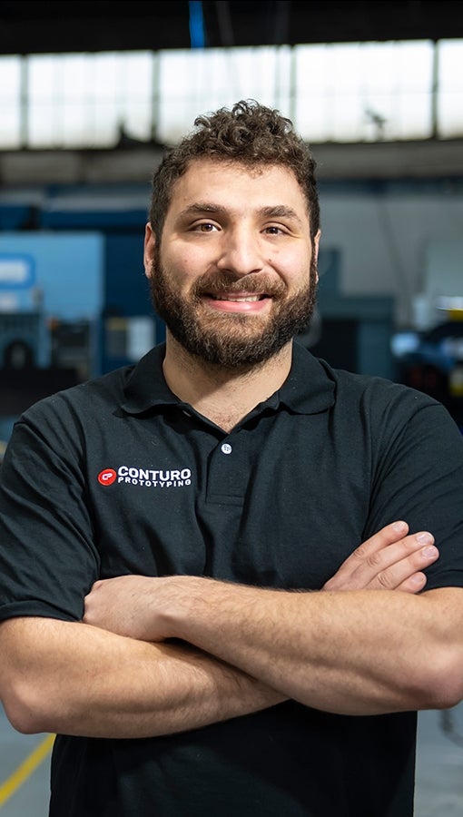 White man with curly hair and beard, wearing navy blue polo that reads "Conturo Prototyping," stands in his company's machine shop, with boxy machinery, old warehouse windows and computer screens in background.