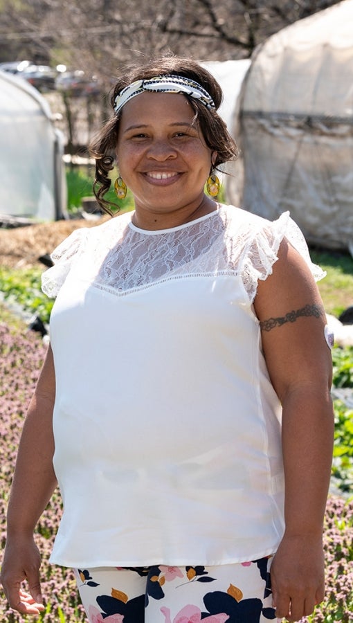 Black woman stands amid rows of plants, including green florets and small pink flowers, and greenhouses.