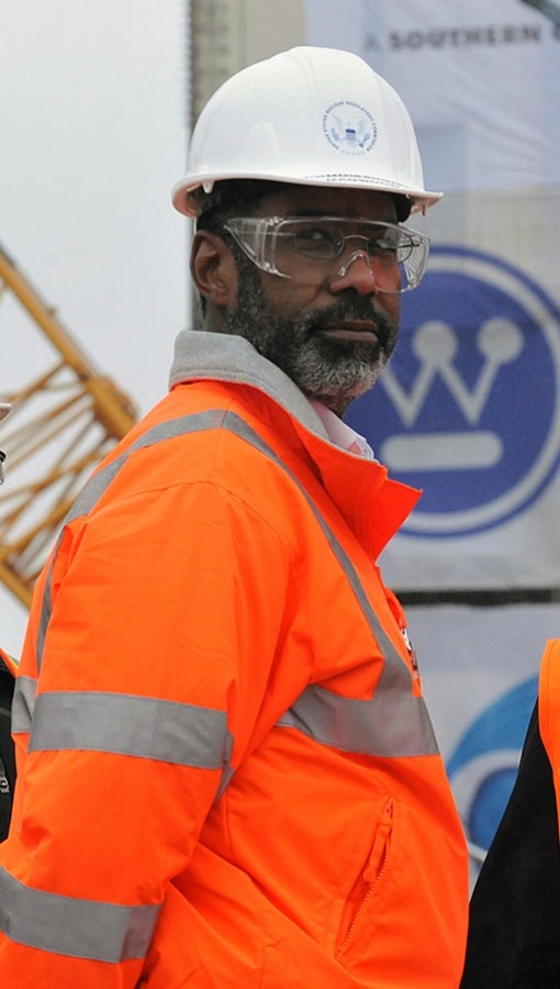 Black man wearing bright orange construction jacket, safety goggles and white hardhat stands in front of crane and Westinghouse logo