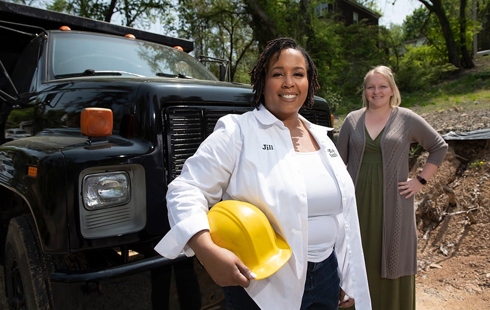 Black woman wearing white button-down shirt and holding yellow hart hat and white woman in long moss-green dress stand in front of large truck.