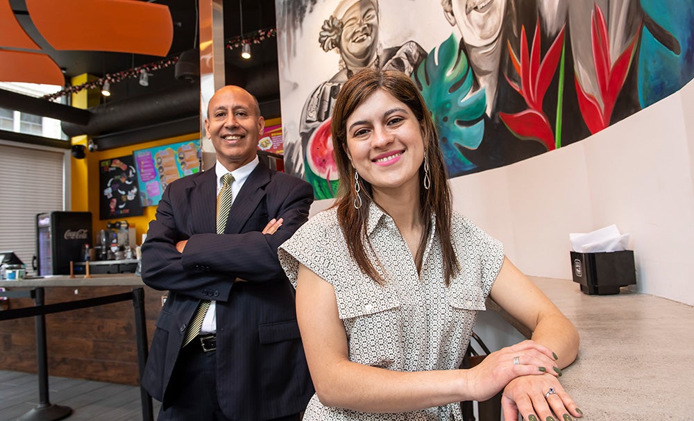 Man in dark suit and gold tie stands with arms crossed; woman with reddish-brown hair wears neutral patterned blouse; both stand in café space in front of colorful mural of women and flowers.