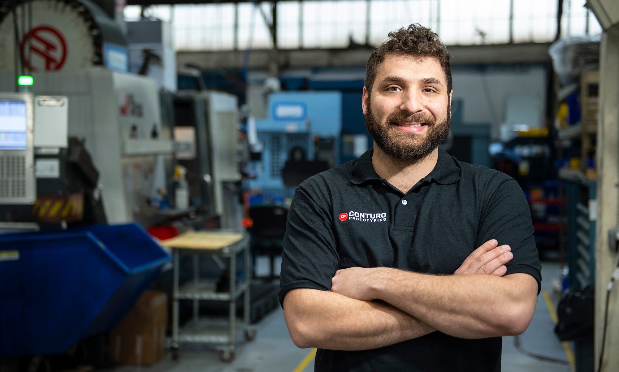 White man with curly hair and beard, wearing navy blue polo that reads "Conturo Prototyping," stands in his company's machine shop, with boxy machinery and computer screens in background.