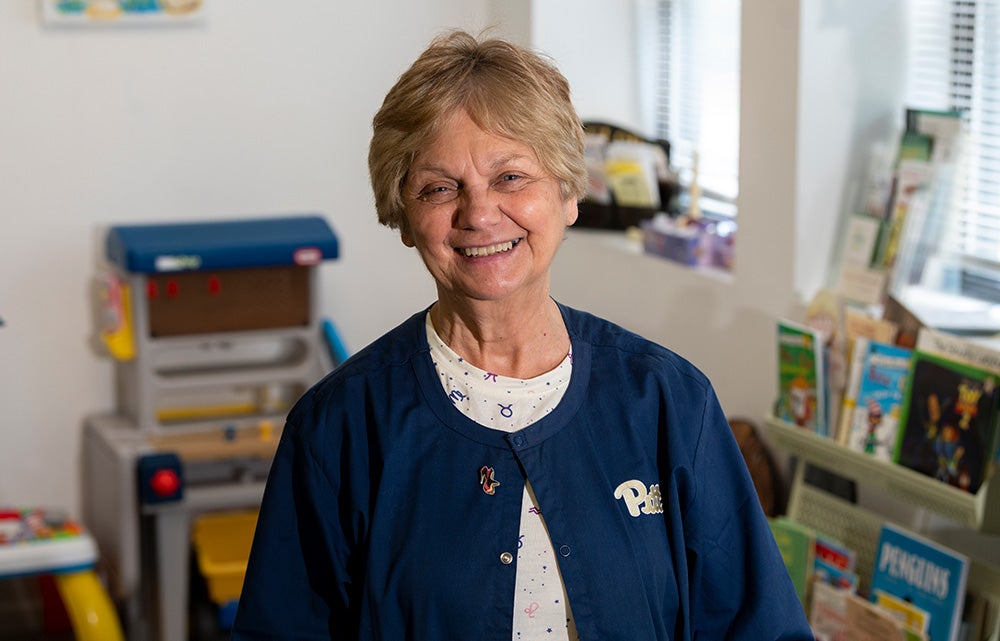 Woman in her '60s wearing a navy blue Pitt fleece stands in room with children's toys and books.