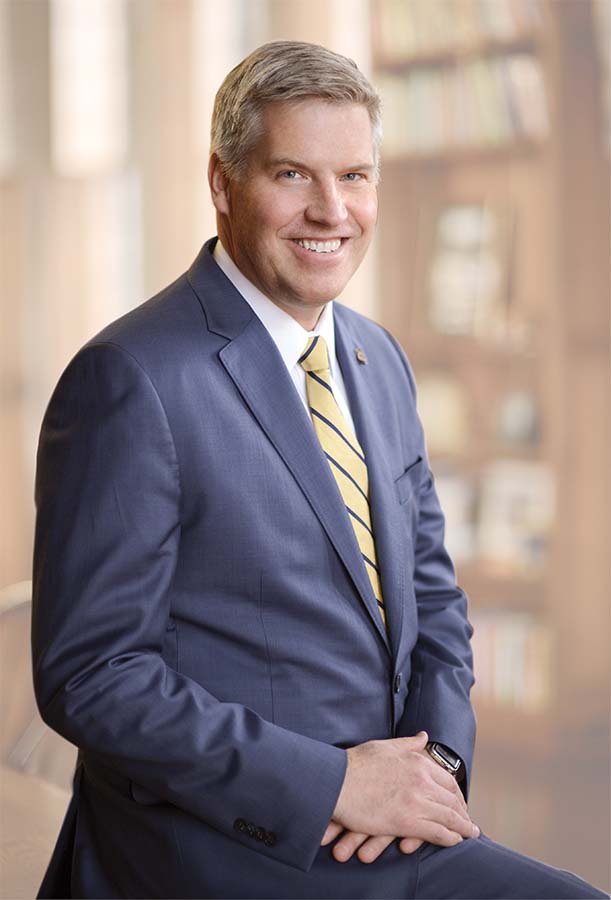 Chancellor Patrick Gallagher in suit in professional portrait, bookshelf and windows blurred in background