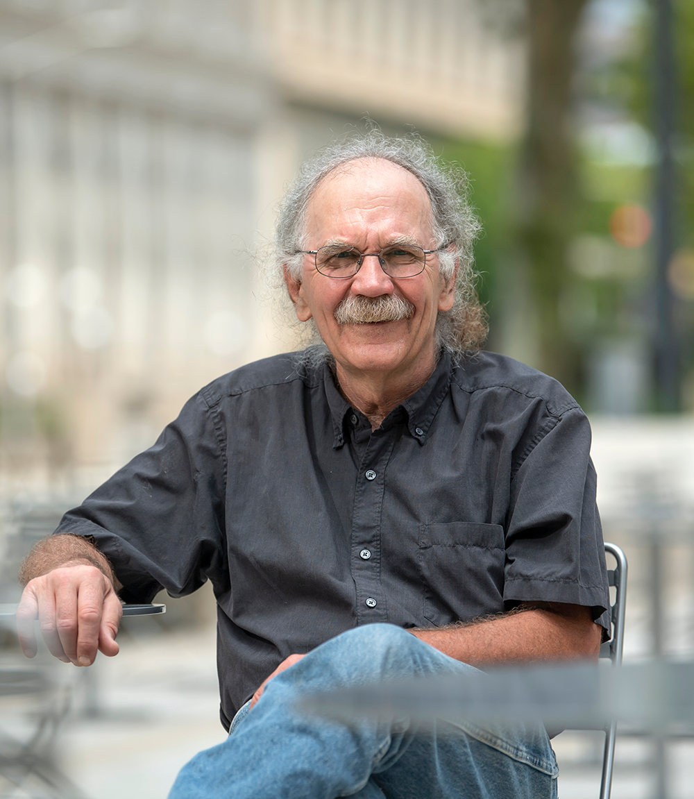 white man with gray curly hair, mustache wearing dark gray button-down and blue jeans