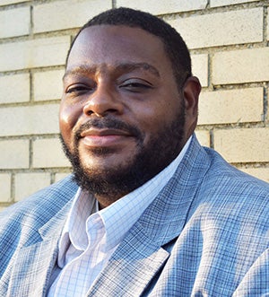 Headshot of Black man in gray plaid suit and striped white shirt