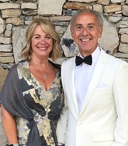 Lisa in a gray floral dress and shawl and John in a white suit with black bowtie in front of stone wall