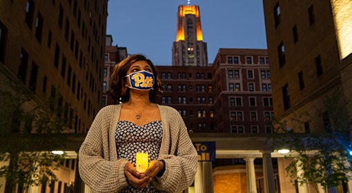 Student wearing tan cardigan and floral dress holds her LED-powered lantern and wears her Pitt script mask in the Quad, the Cathedral lights on in the background