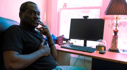 Black man in black t-shirt and turquoise slacks sits at a desk in front of a window, with a computer monitor, keyboard and table lap 