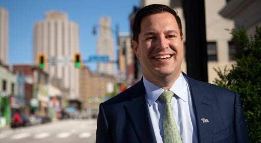 Main with dark hair, navy blue suit, light green tie and script Pitt pin on lapel stands sidewalk in Oakland, with Forbes Ave., Towers and Cathedral of learning visible in background