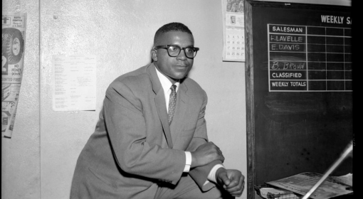 Young black man in suit and tie leans on knee, leg propped up, wears dark rimmed glasses in an interior space with chalkboard