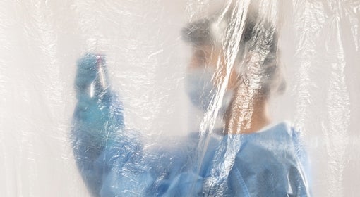 woman wearing blue protective gear and face mask holding injection in front of her while standing behind clear sheet of plastic