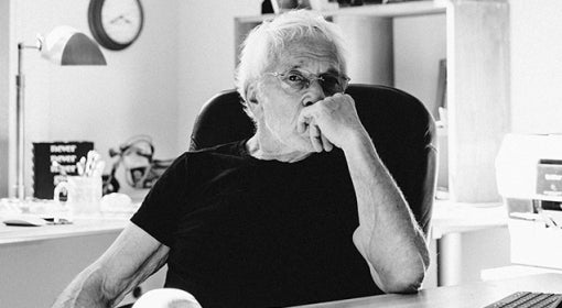 black and white photo of Lee Gutkind, in short-sleeved black t-shirt, sitting behind desk in his office