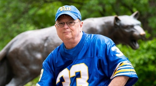 man wears blue and gold 33 football jersey and Pitt script hat in front of panther statue