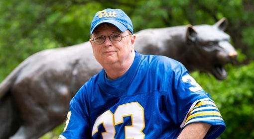 man wears blue and gold 33 football jersey and Pitt script hat in front of panther statue