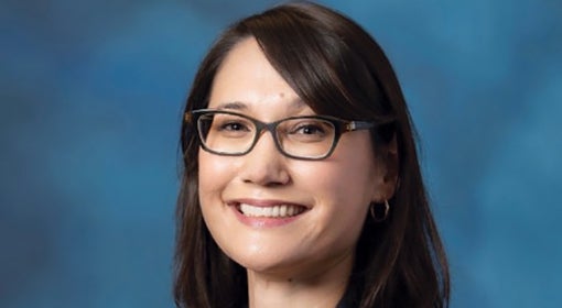 Woman with dark straight hair, and dark-rimmed glasses smiles at camera in front of teal background