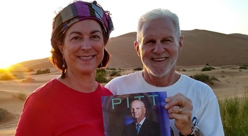 woman in red shirt and headband and man in white University of Pittsburgh T-shirt hold Pitt Magazine Fall 2019 issue as the sun goes down on sandy desert landscape