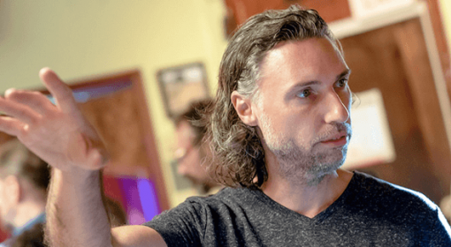 Man with shoulder-length silver hair, wearing dark gray t-shirt, holds up hand while talking with someone