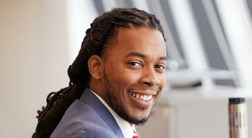 professional portrait of young Black man in blue-grey suit, white button-down shirt, red tie. He is smiling at camera.