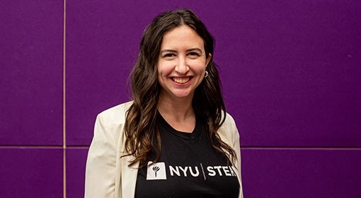 Sara Klein stands in a black NYU shirt and white jacket against a purple wall