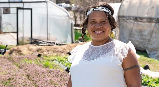 Black woman stands amid rows of plants, including green florets and small pink flowers, and greenhouses.