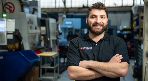 White man with curly hair and beard, wearing navy blue polo that reads "Conturo Prototyping," stands in his company's machine shop, with boxy machinery and computer screens in background.