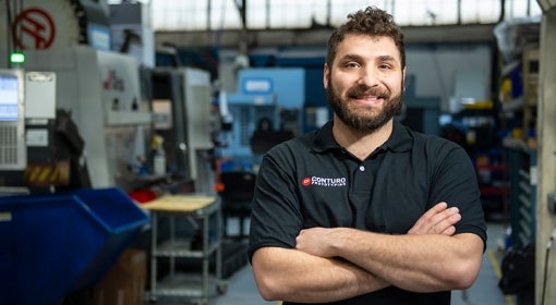 White man with curly hair and beard, wearing navy blue polo that reads "Conturo Prototyping," stands in his company's machine shop, with boxy machinery and computer screens in background.