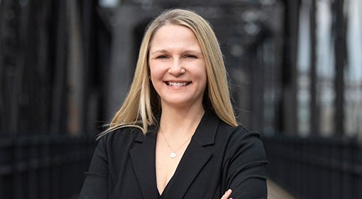 Woman in black blazer and long blond hair smiles at camera and stands on a black, industrial-style bridge