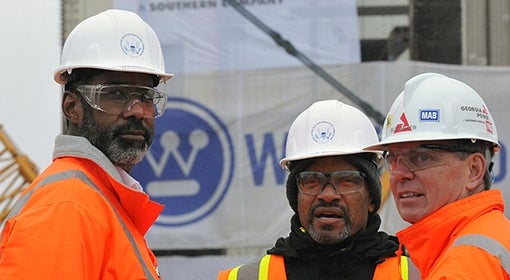 Three men wear white hard hats and bright orange construction vests/jackets, with crane and sign for and "Westinghouse" in the background