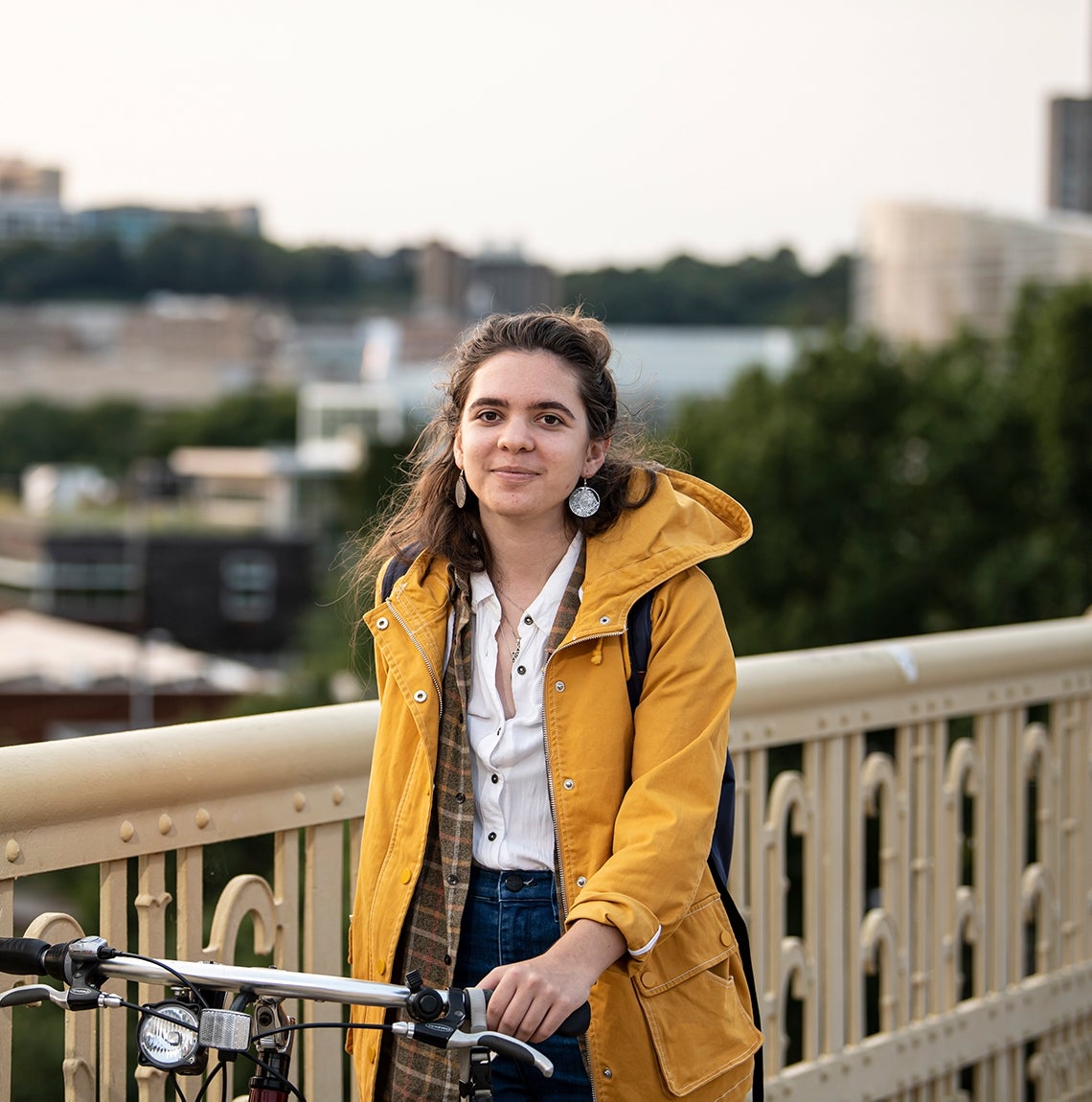 Avocet Greenwell, wearing a yellow coat, plaid shirt, a backpack, dangle earrings and jeans, stands on Schenley Bridge with her bike