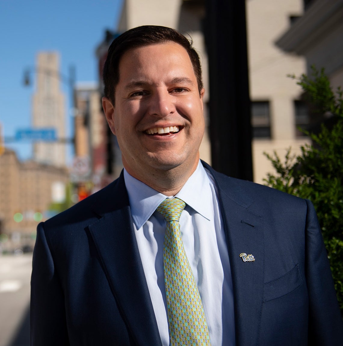 Main with dark hair, navy blue suit, light green tie and script Pitt pin on lapel stands sidewalk in Oakland, with Forbes Ave. and the Cathedral of learning visible in background
