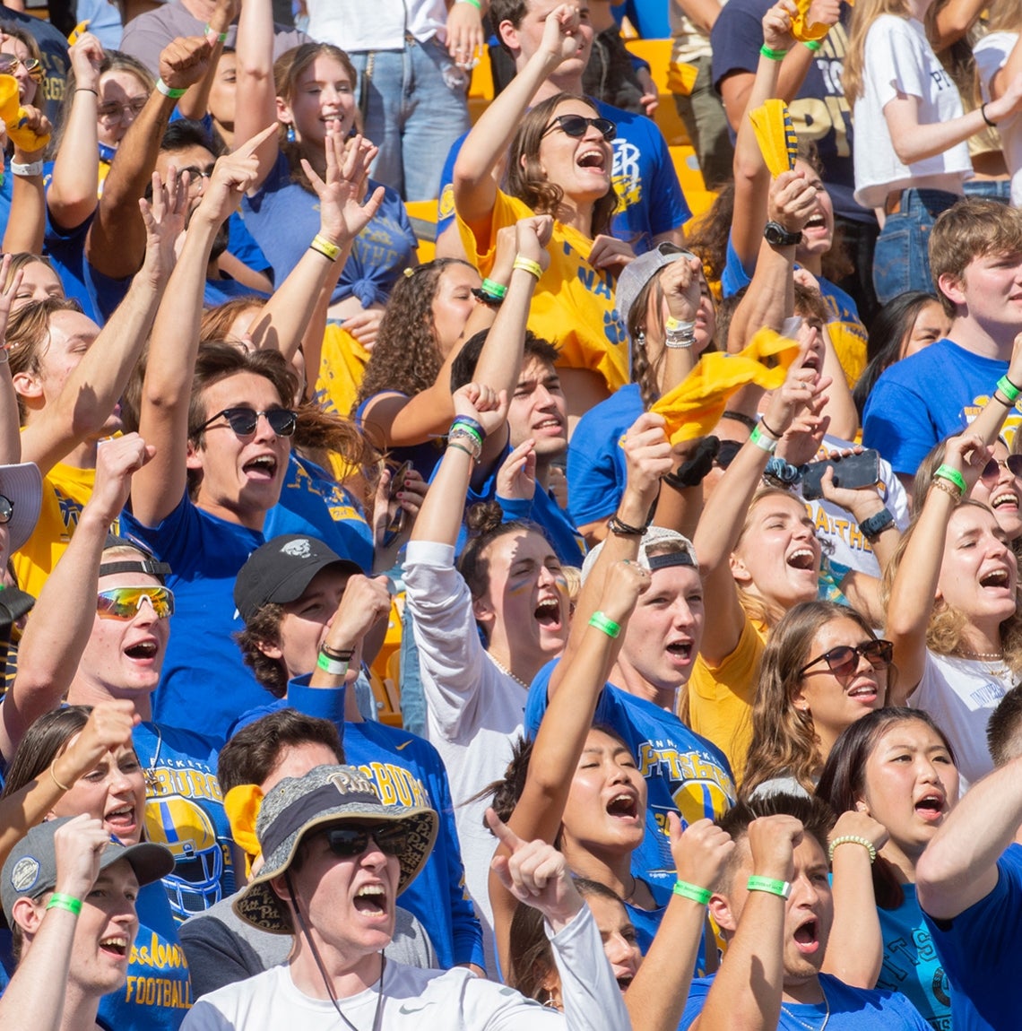 crowd of students wearing blue and gold, arms raised, yelling and standing in stadium seating