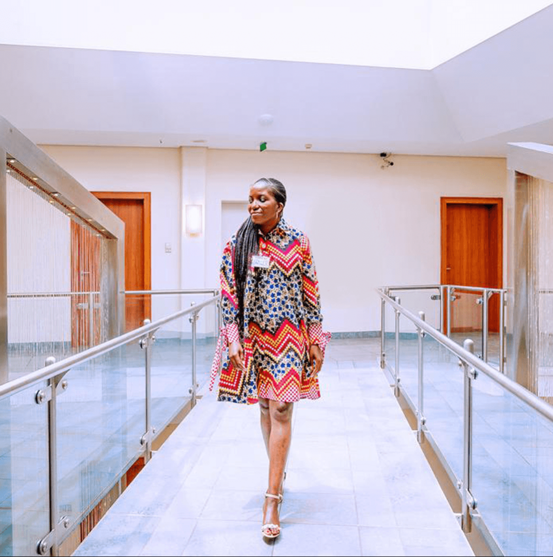 Black African woman wearing print dress and walking in glass-paned, sunlit hallway