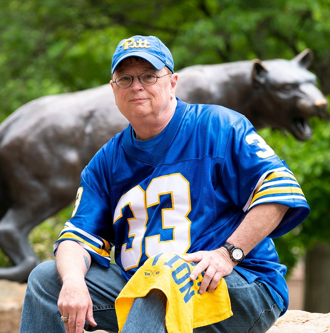 man wears blue and gold 33 football jersey and Pitt script hat in front of panther statue