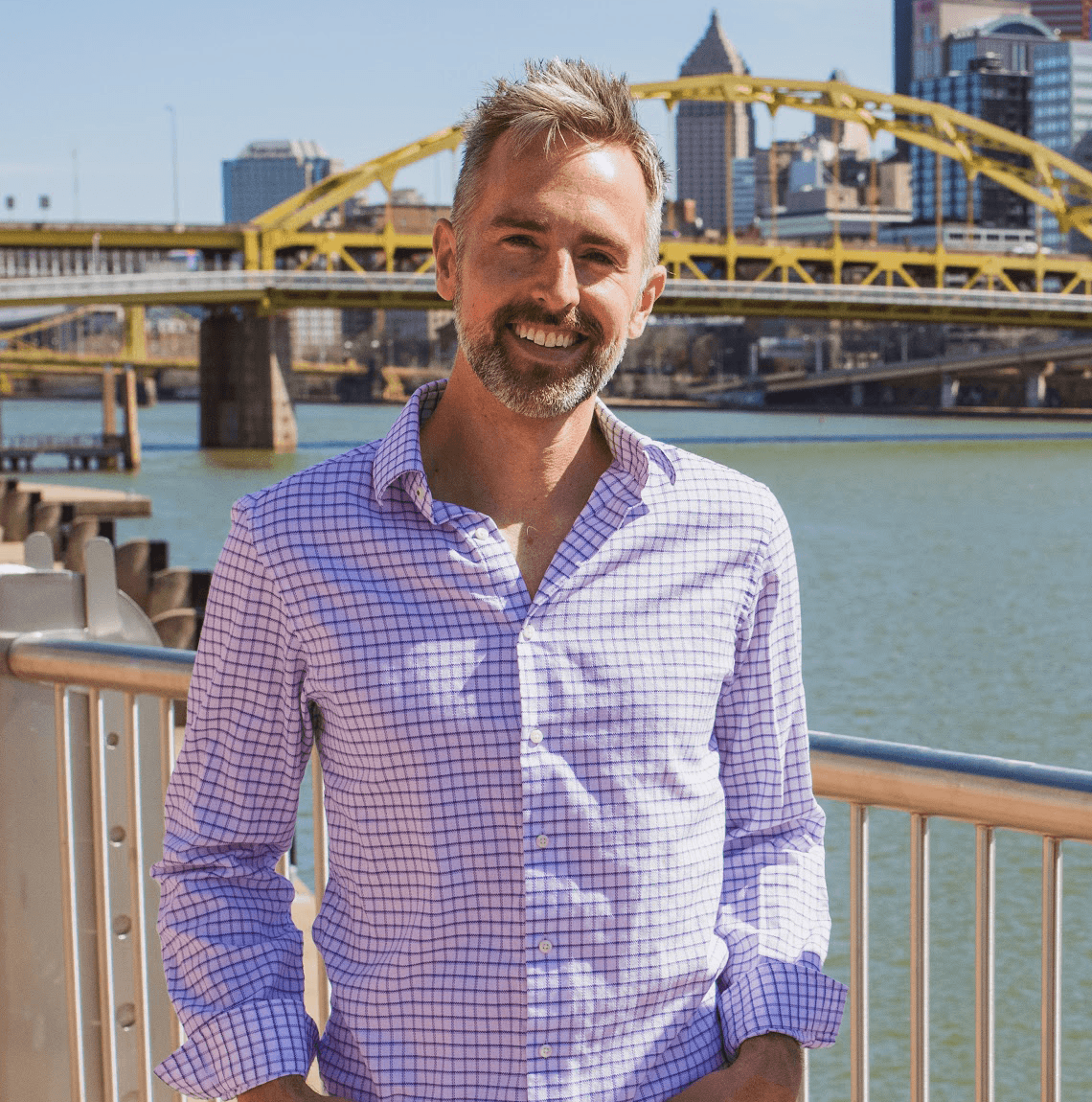 photo of White man with gray hair and beard in button-down shirt stands in front of river, bridge and cityscape 