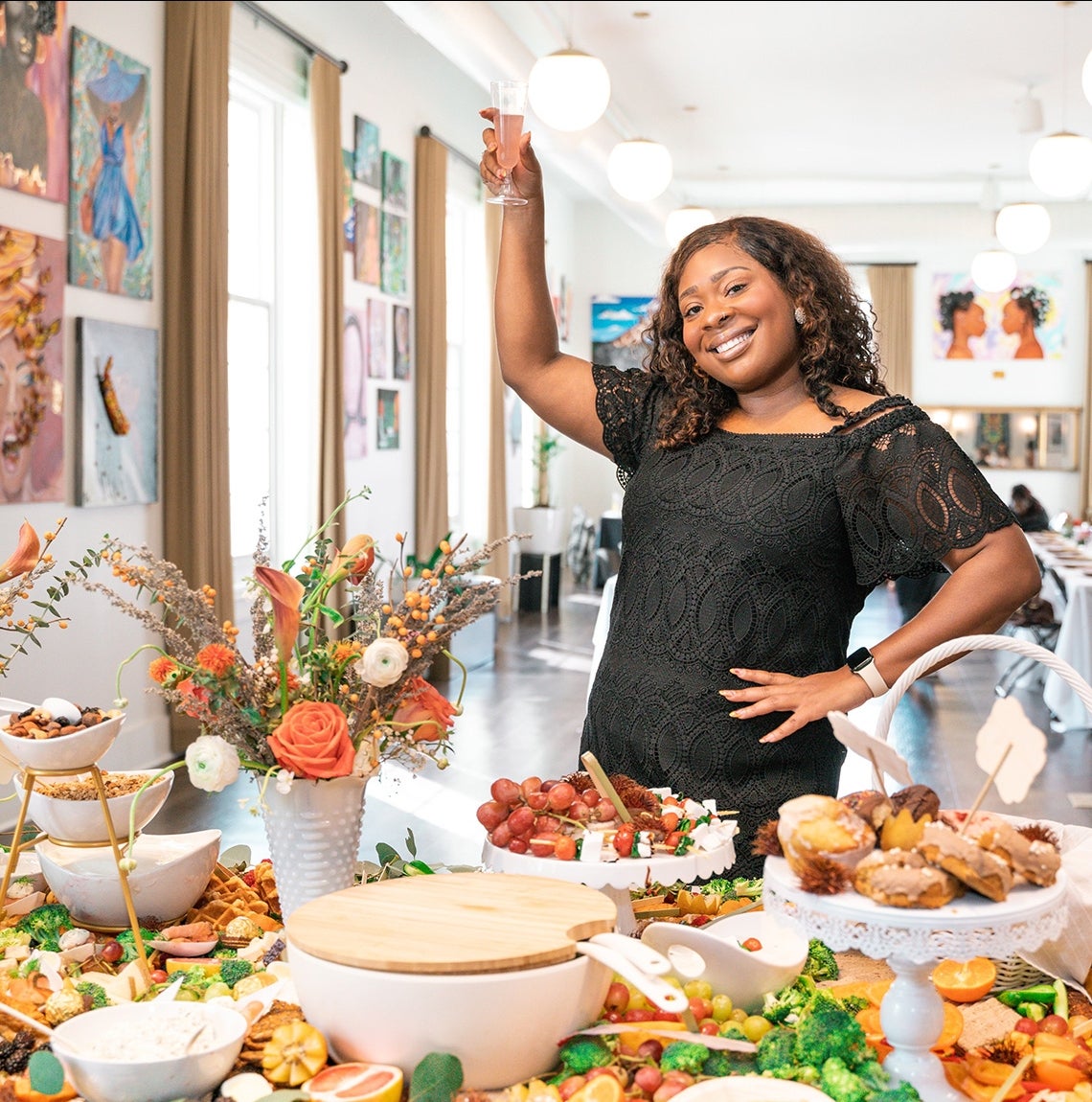 A person in a black dress holds up a glass behind a table of food and flowers