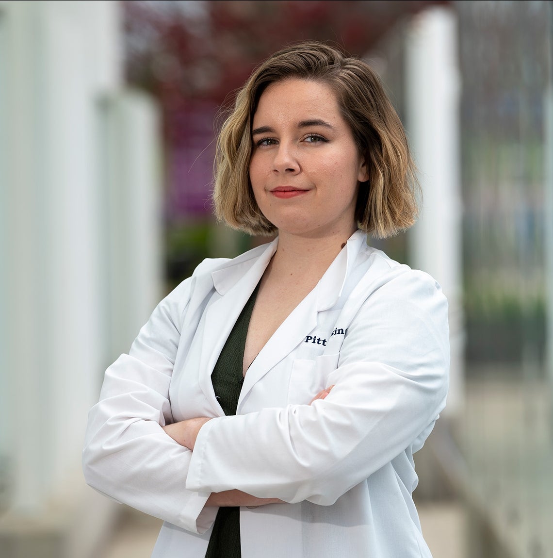 Young woman with short, light-brown hair and white coat with lapels stands with arms crossed in front of glass panels on sidewalk.