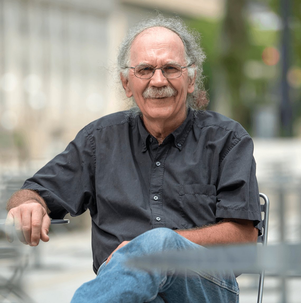 white man with gray curly hair, mustache wearing dark gray button-down and blue jeans