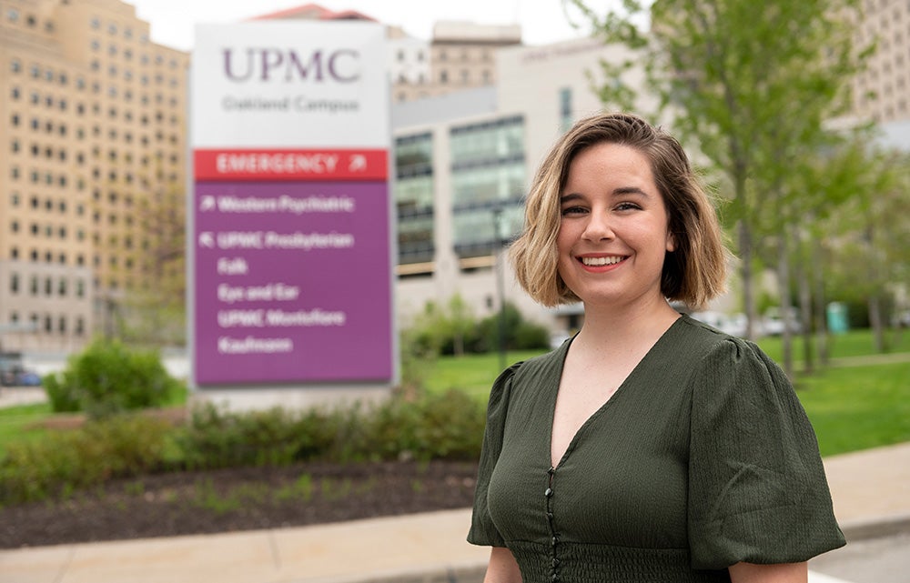 Young woman with brown-blond bob wearing moss-green dress stands in front of UPMC Presbyterian Oakland Campus and hospital buildings in background